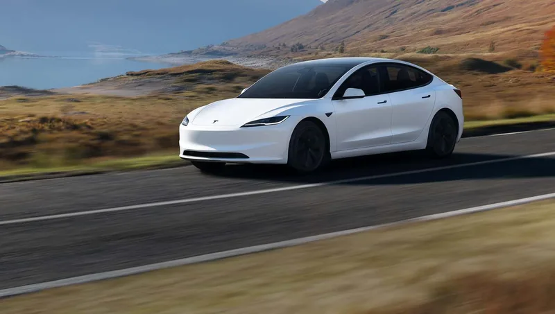 White Tesla Model 3 sedan driving on a mountain road above a lake.